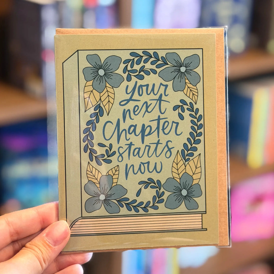 Card with floral design and text held by a hand in a blurred bookstore setting. Text reads 'Your next chapter starts now'