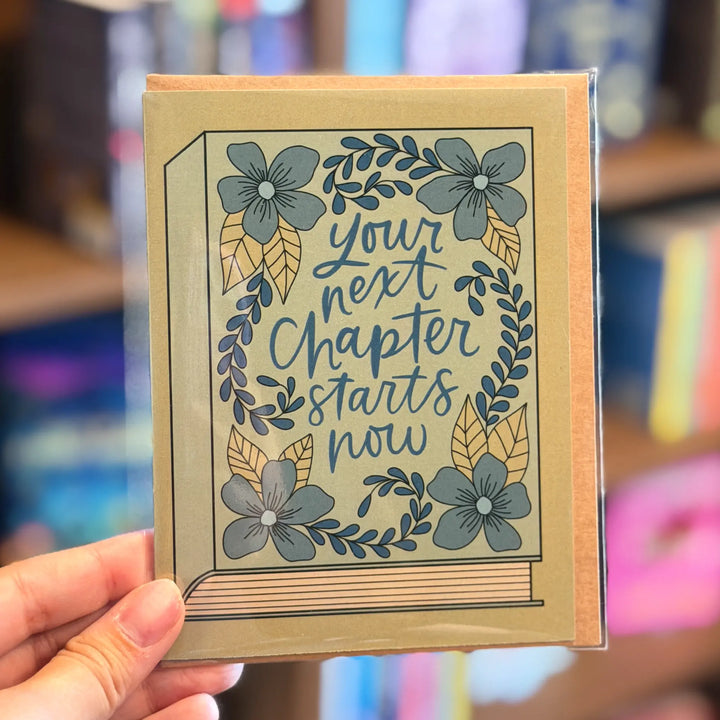 Card with floral design and text held by a hand in a blurred bookstore setting. Text reads &