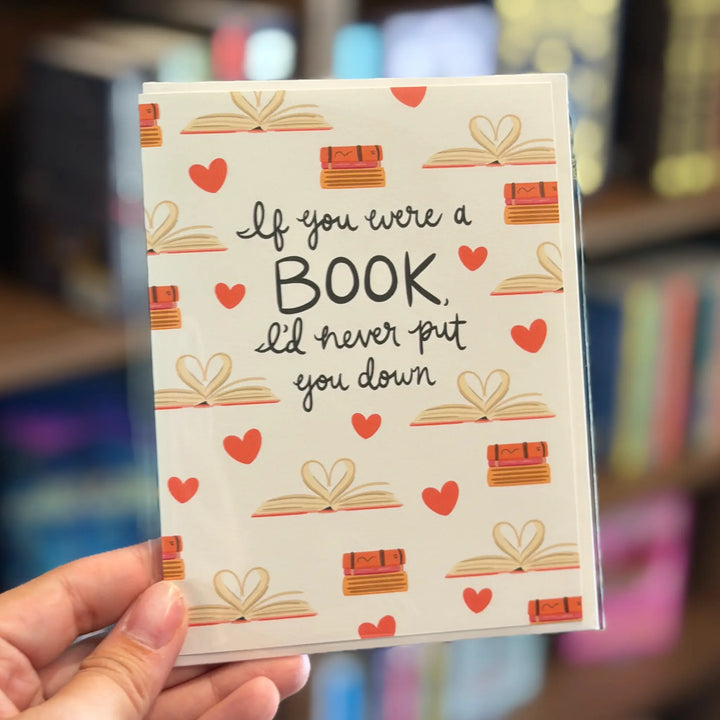 Card with book-themed design and text held in front of a blurred bookshelf background. Text reads &