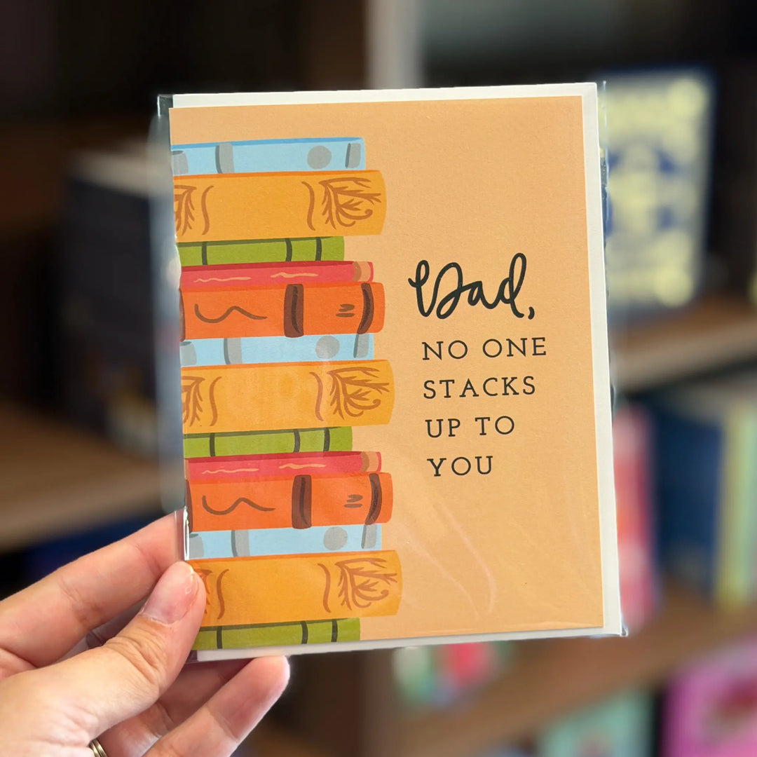 Card with book illustration and text held by a hand, blurred background. Text reads 'Dad, no one stacks up to you'