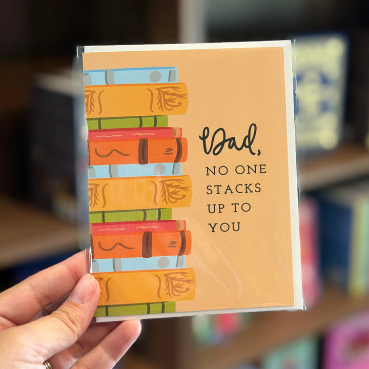 Card with book illustration and text held by a hand, blurred background. Text reads &