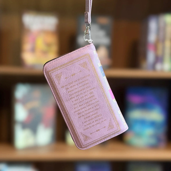 Pink book-shaped wallet with text and decorative elements, hanging against a blurred bookshelf background.