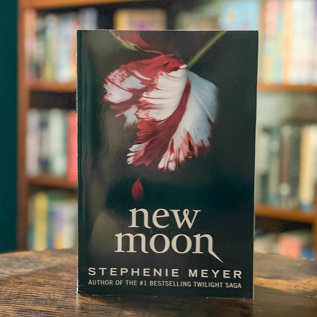 Book titled 'New Moon' by Stephenie Meyer with a flower design on the cover, placed on a wooden surface with a blurred bookshelf background.