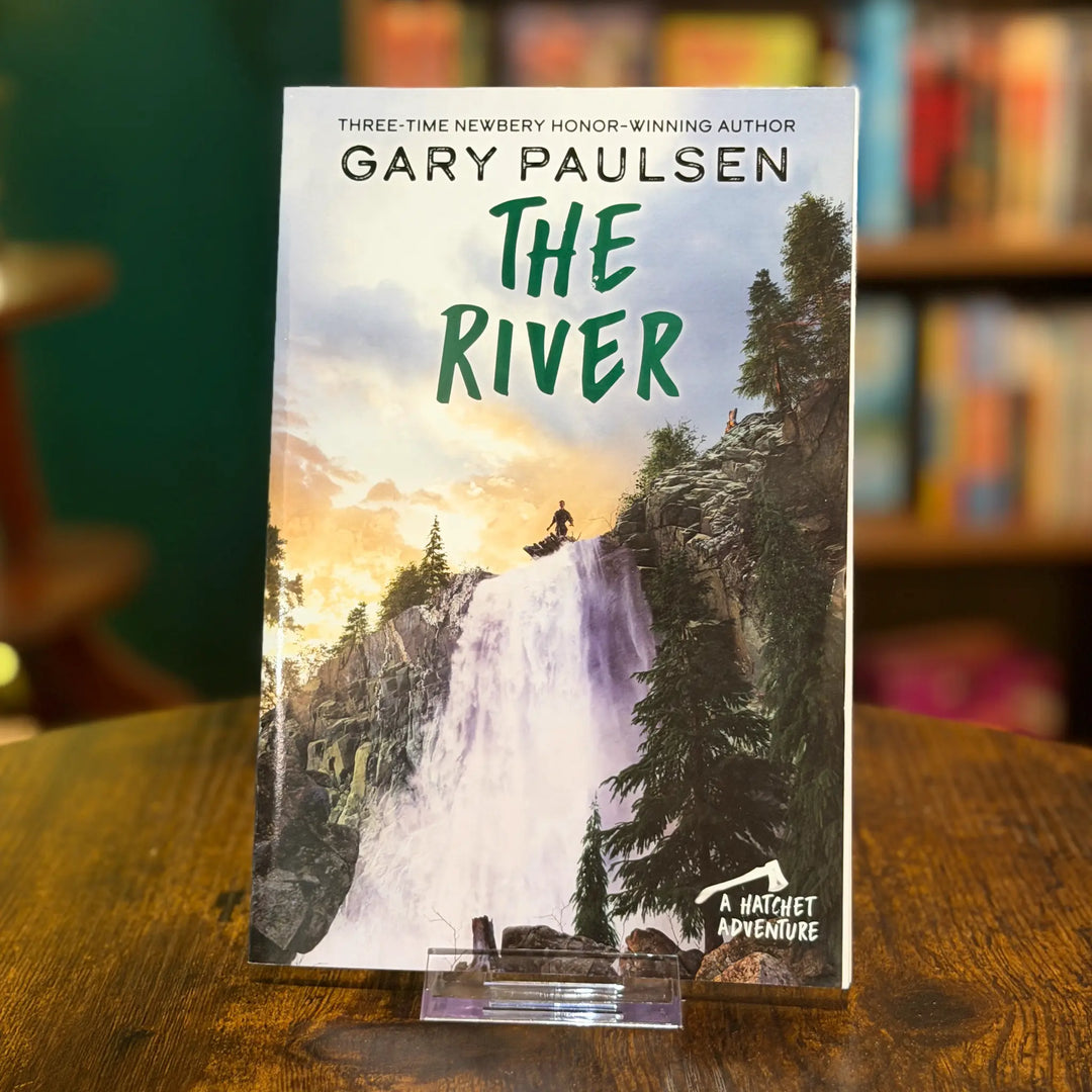 Book titled 'The River' by Gary Paulsen on a wooden surface with a blurred bookshelf background.