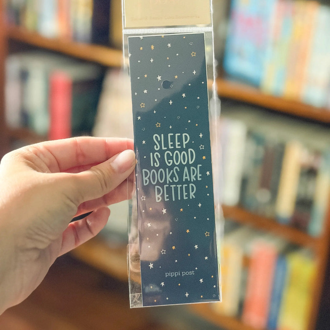 Hand holding a bookmark with 'Sleep is good, books are better' text in a library setting