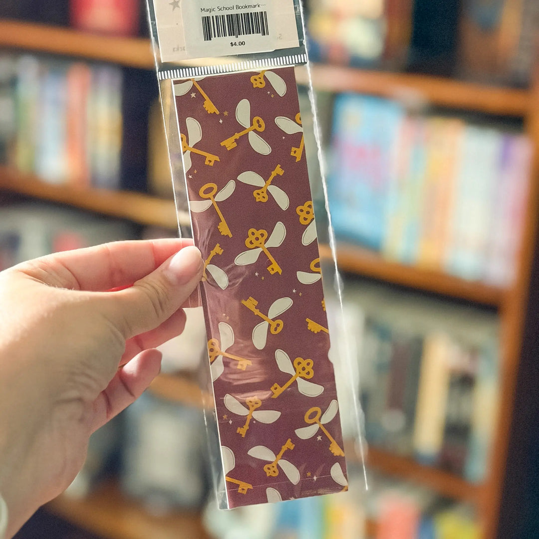 Packaged bookmarks with dragonfly pattern held by a hand in front of a bookshelf.
