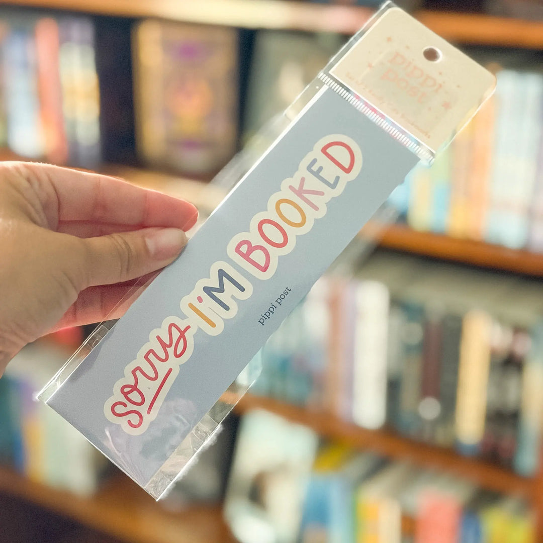 Hand holding a bookplate with 'Sorry I'm Booked' text against a blurred bookshelf background
