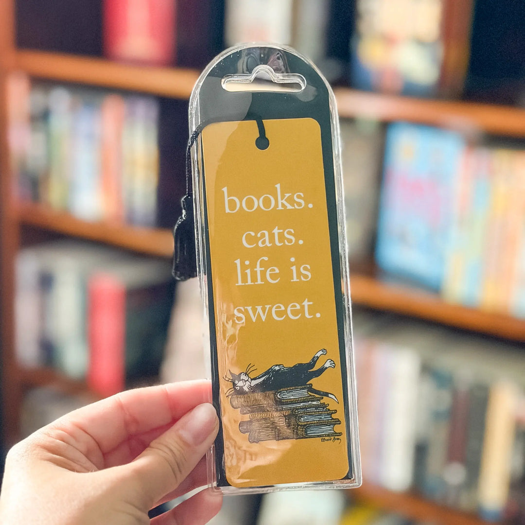 Hand holding a yellow bookmark with text and a cat illustration in front of a bookshelf.