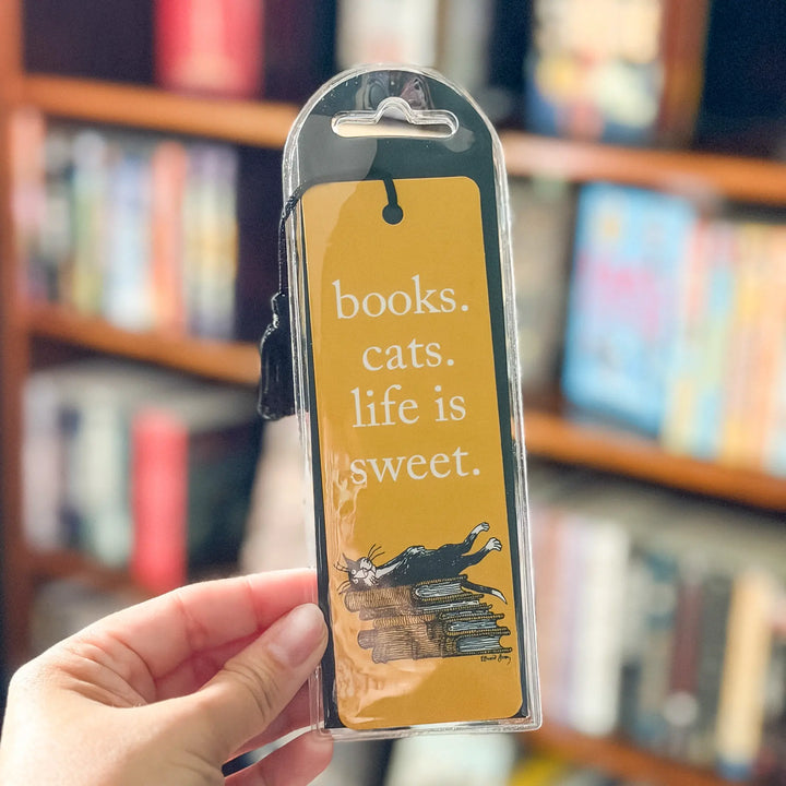 Hand holding a yellow bookmark with text and a cat illustration in front of a bookshelf.