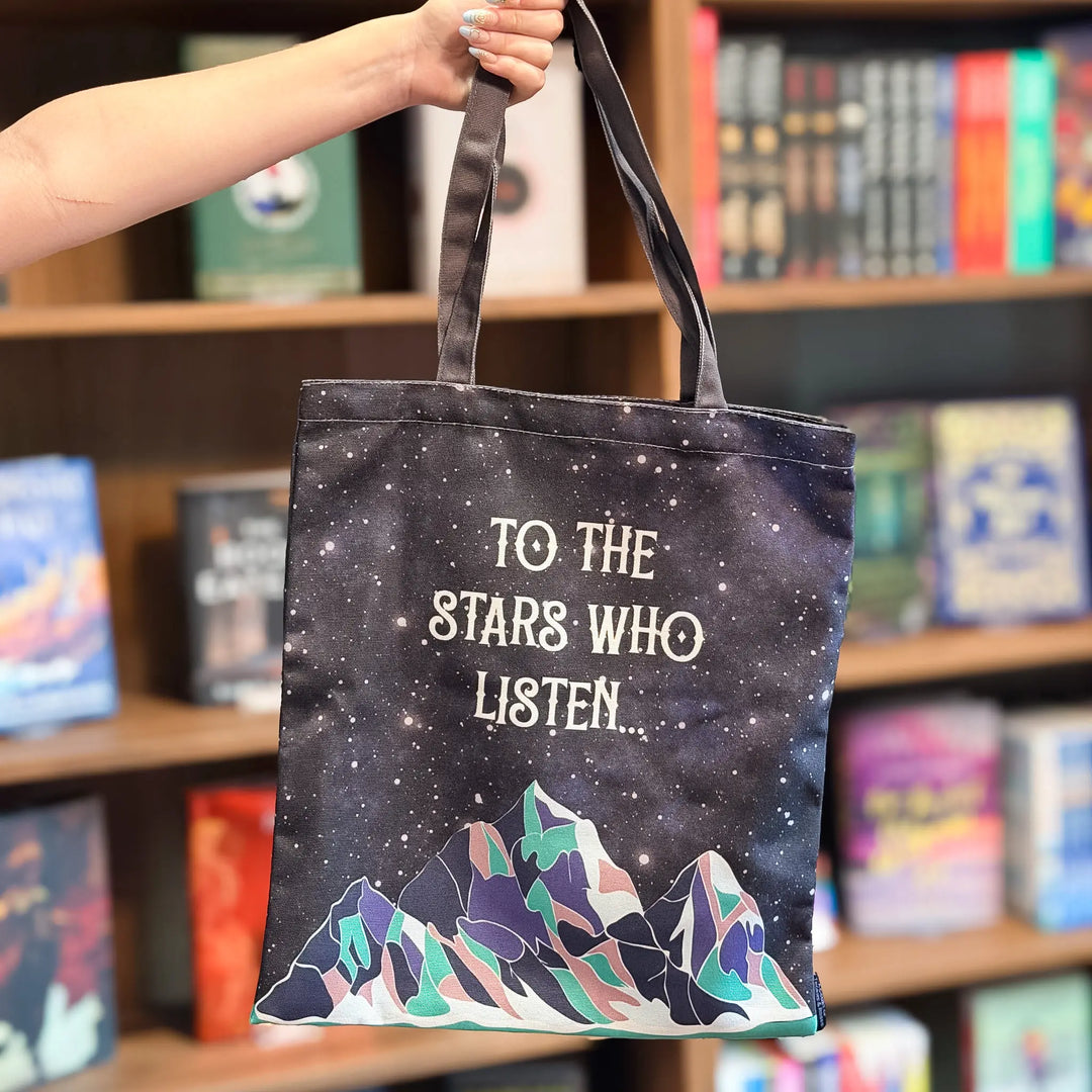 Tote bag with 'To the Stars Who Listen' text and mountain design held in front of a bookshelf.
