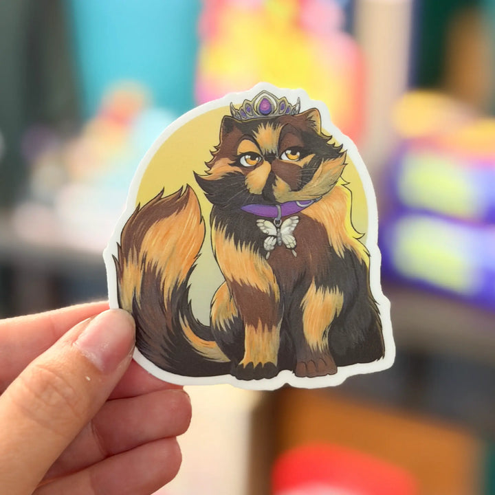 Sticker of a cat with a crown held by a hand against a colorful blurred background