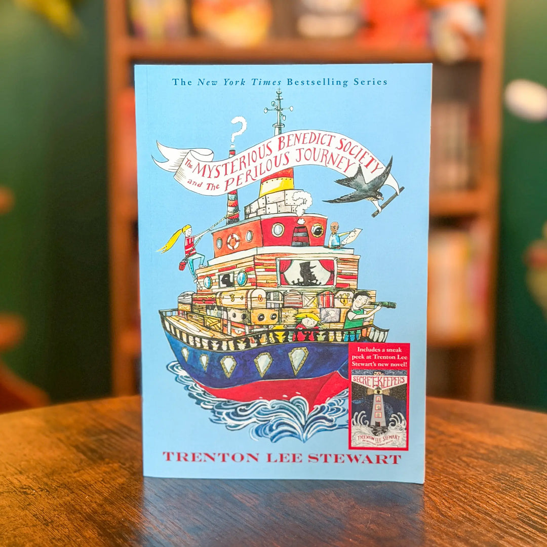 Book cover of 'The Mysterious Benedict Society and the Perilous Journey' by Trenton Lee Stewart on a wooden surface with a blurred bookshelf background.