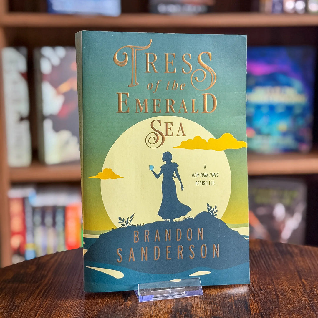 Book titled 'Tress of the Emerald Sea' by Brandon Sanderson on a wooden surface with a blurred bookshelf background.