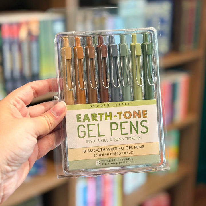 Pack of Earth-tone gel pens held by a hand with a blurred bookshelf background