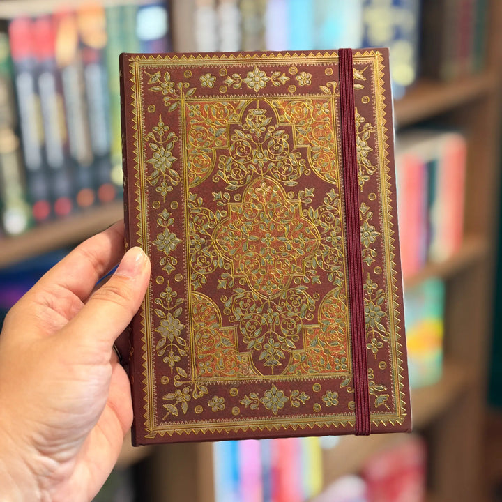 Hand holding a ornate red and gold notebook with bookshelf background