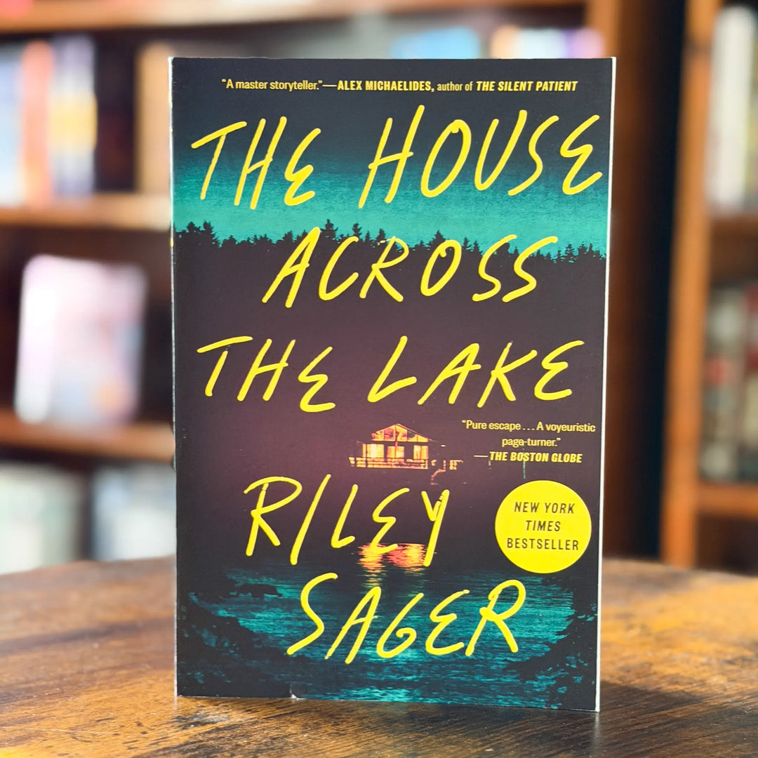 Book titled 'The House Across the Lake' by Riley Sager on a wooden surface with a blurred bookshelf background.