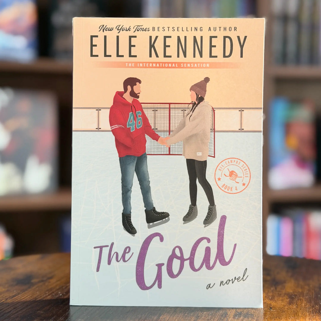 Book cover of 'The Goal' by Elle Kennedy on a wooden surface with blurred bookshelf background