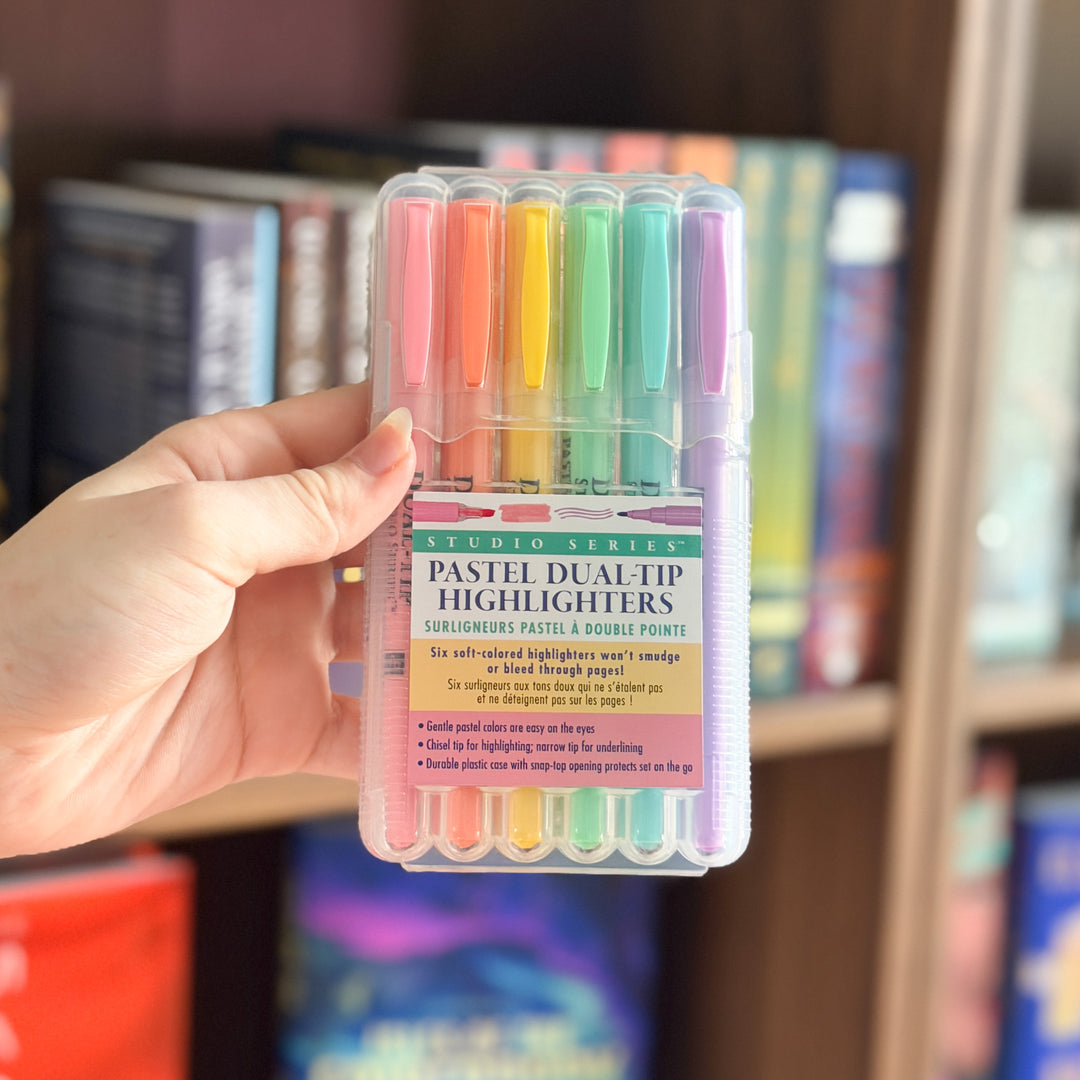 A hand holds a clear plastic case of six pastel dual-tip highlighters in front of a blurred bookshelf background. The highlighters come in pink, orange, yellow, green, blue, and purple, with a label reading &