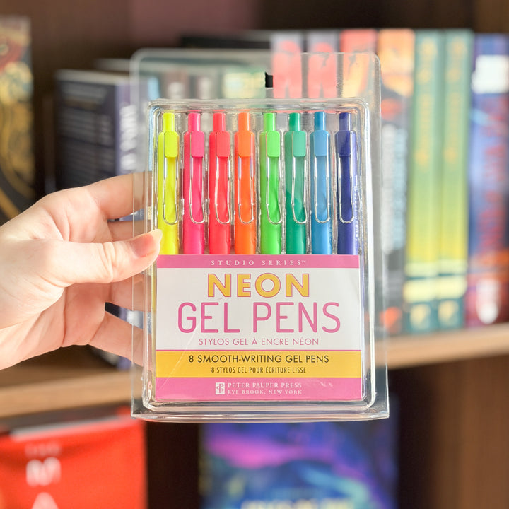 A hand holds a clear plastic case containing eight Studio Series Neon Gel Pens in assorted bright colors, with a blurred bookshelf background.