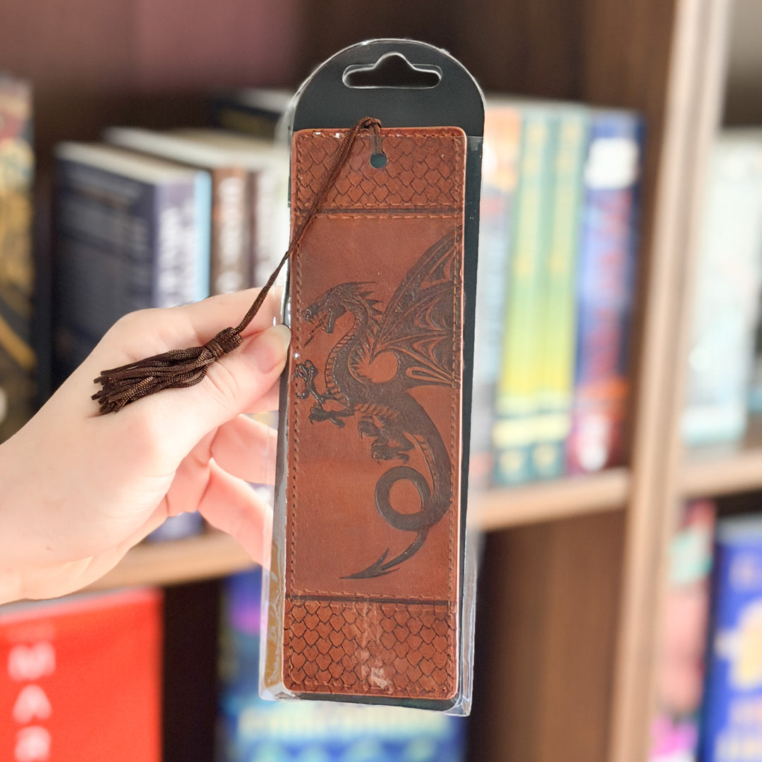 A hand holds a brown leather bookmark featuring an embossed dragon design and a tassel, still in its clear plastic packaging with a black header card, with a blurred bookshelf in the background.