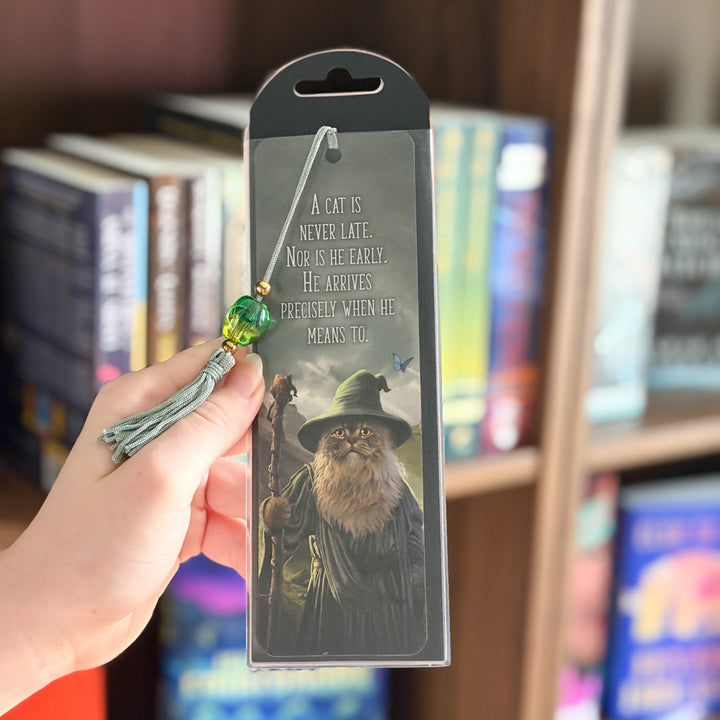 A hand holds a packaged bookmark featuring a wizard cat illustration and the quote &