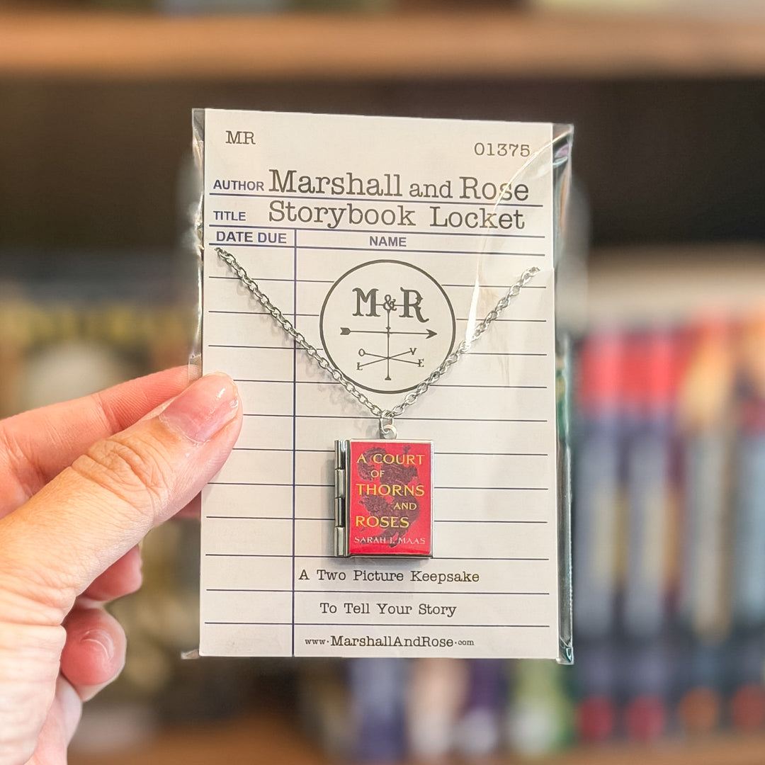 Necklace with 'M' and 'R' design on a book-shaped pendant, displayed on a library card.