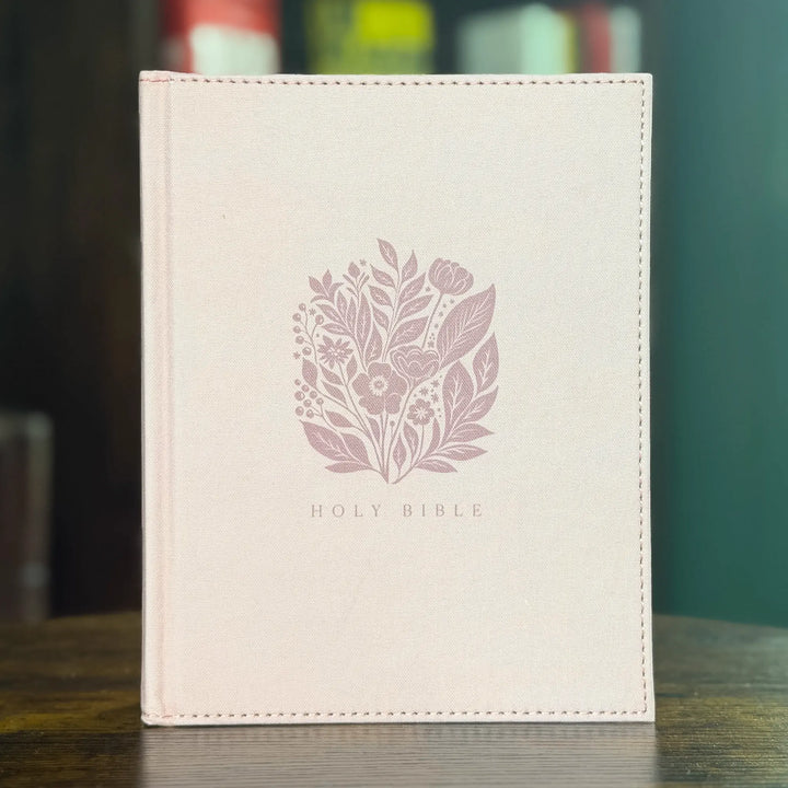 A light pink cloth-bound Holy Bible standing upright on a wooden surface, featuring a muted pink floral illustration and the words &