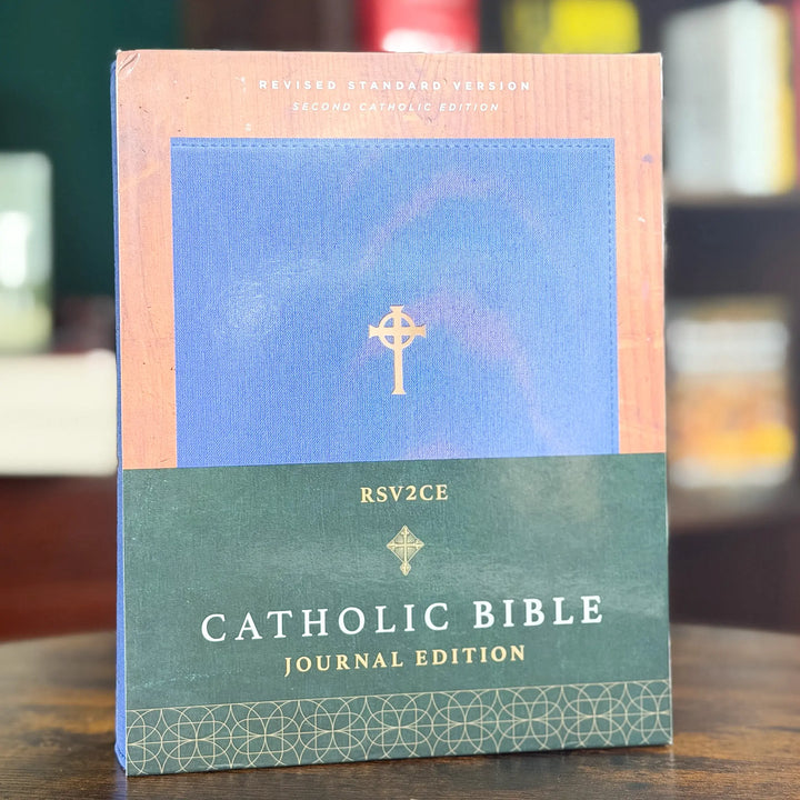 A Catholic Bible Journal Edition book standing upright on a wooden surface, with a blue upper section, peach border, dark green lower section, and gold text and cross details.