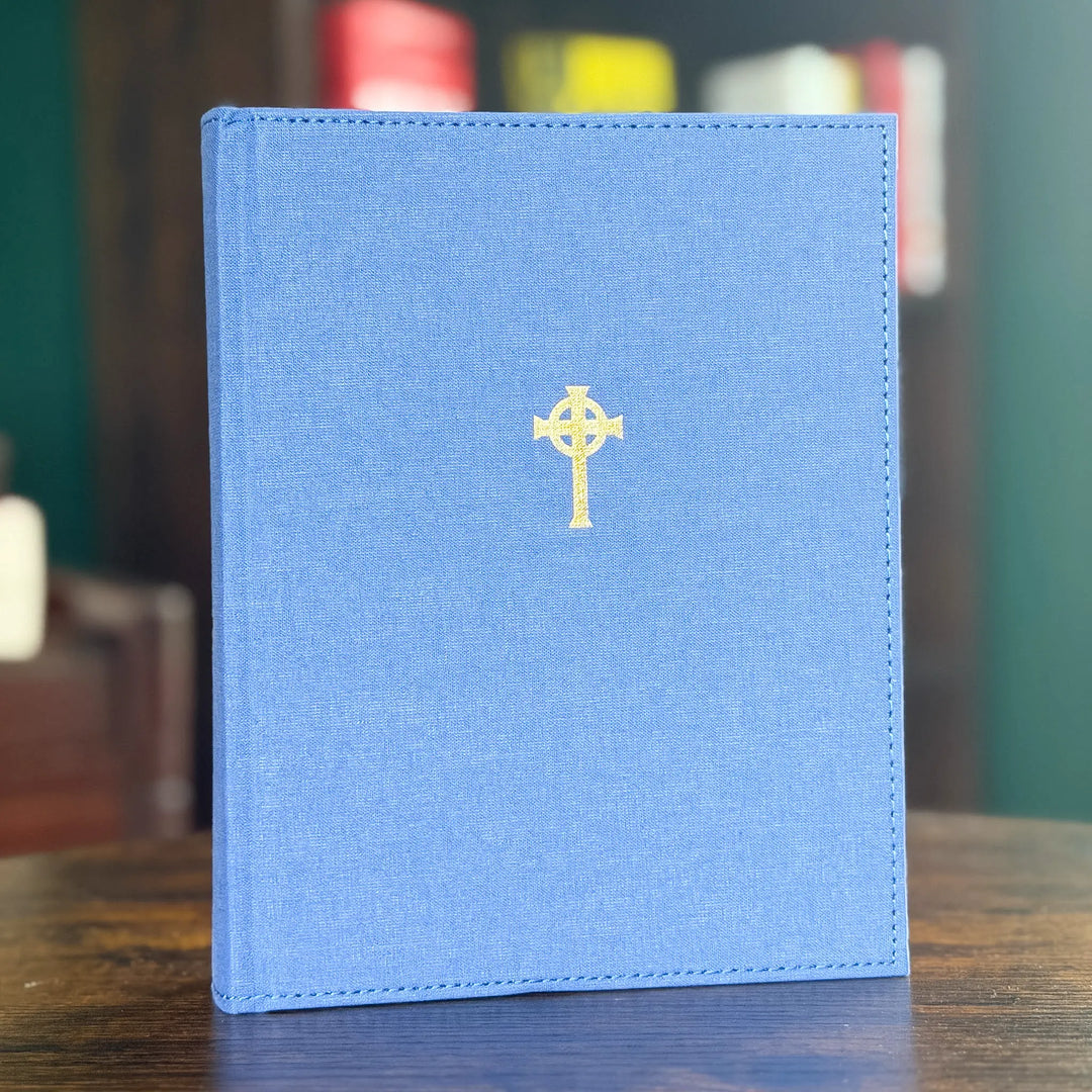 A blue linen-covered book standing upright on a wooden surface, featuring a small gold Celtic cross emblem centered on the front cover.