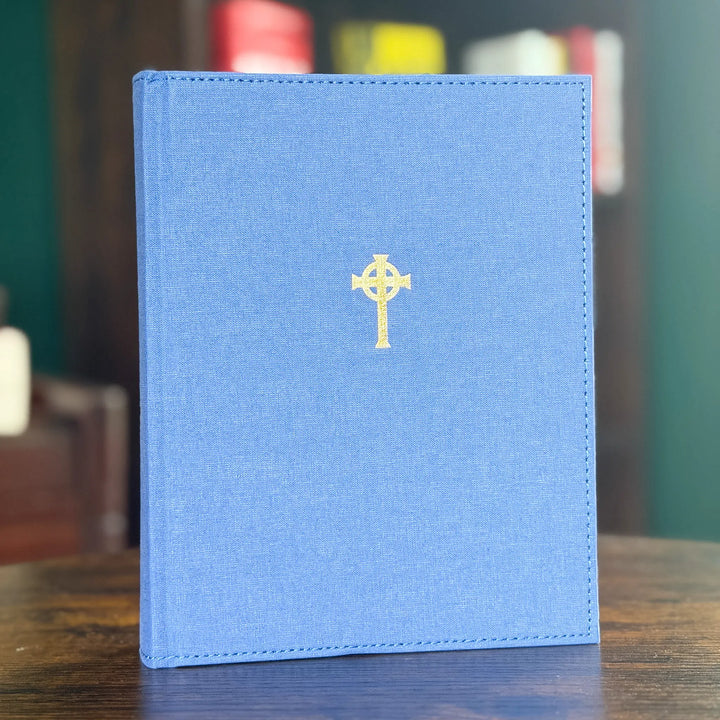 A blue linen-covered book standing upright on a wooden surface, featuring a small gold Celtic cross emblem centered on the front cover.