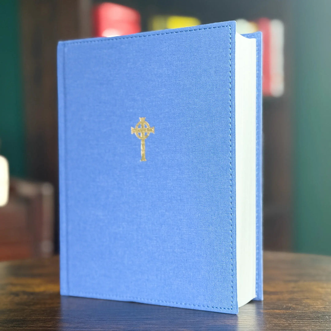 A blue hardcover book standing upright on a wooden table, with a small gold cross emblem centered on the front cover and a blurred indoor background.