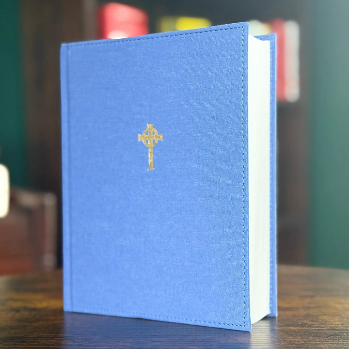 A blue hardcover book standing upright on a wooden table, with a small gold cross emblem centered on the front cover and a blurred indoor background.
