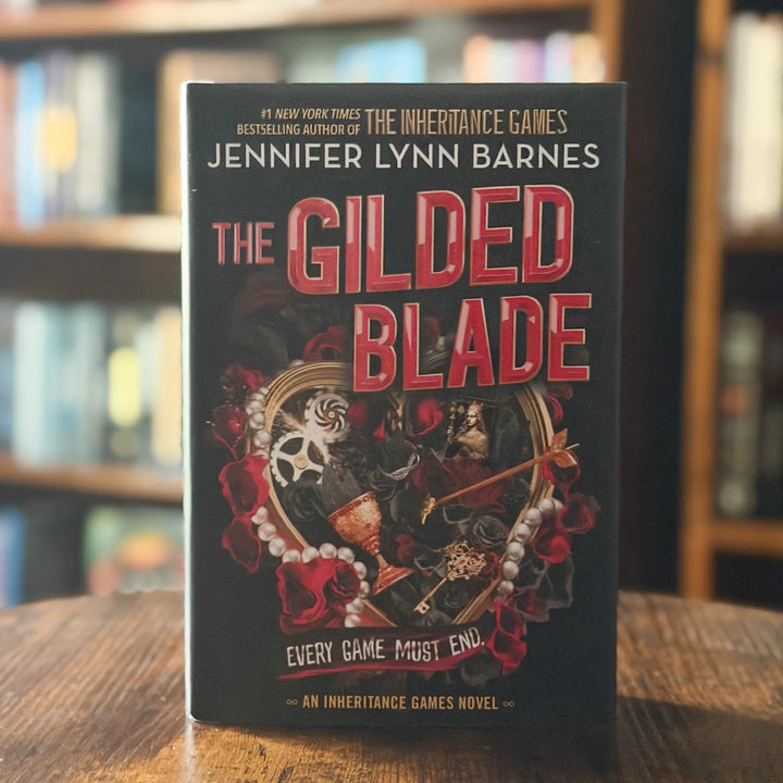 The Gilded Blade book by Jennifer Lynn Barnes standing upright on a wooden table with blurred bookshelves in the background.