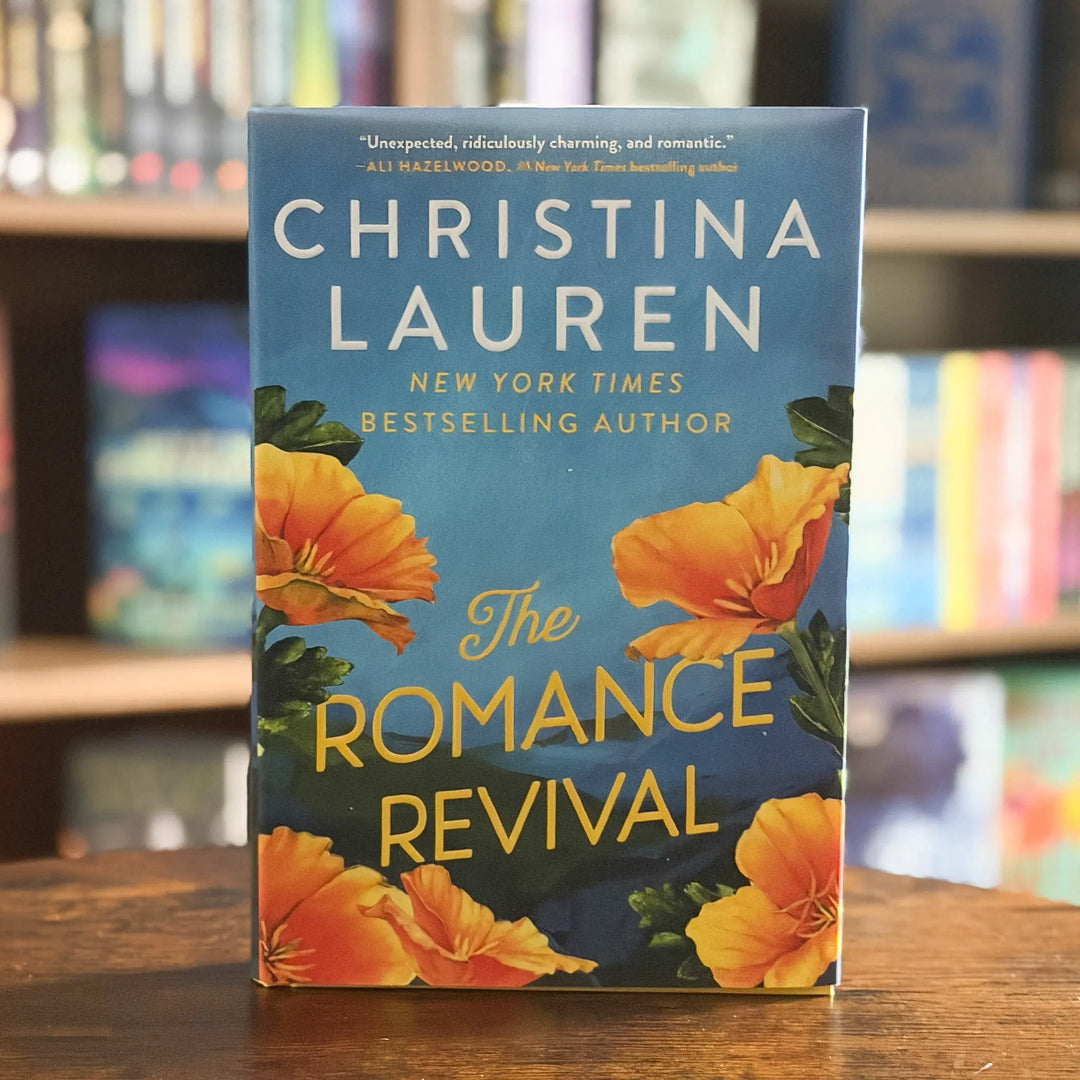 The Romance Revival paperback book by Christina Lauren, featuring a blue cover with yellow-orange flowers, standing on a wooden table with a blurred bookshelf background.