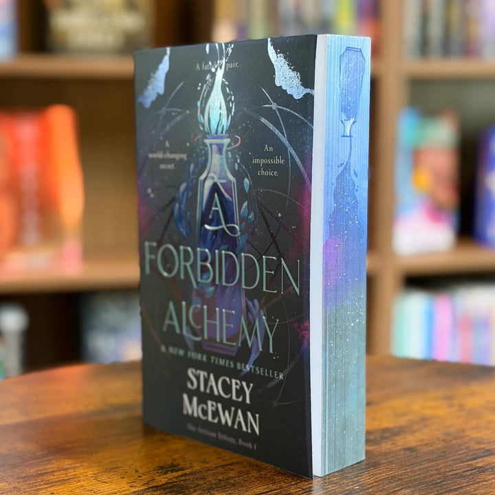 A hardcover book titled "Forbidden Alchemy" by Stacey McEwan stands upright on a wooden table with blurred bookshelves in the background. The cover is predominantly black with glowing blue and purple artwork and white title text.