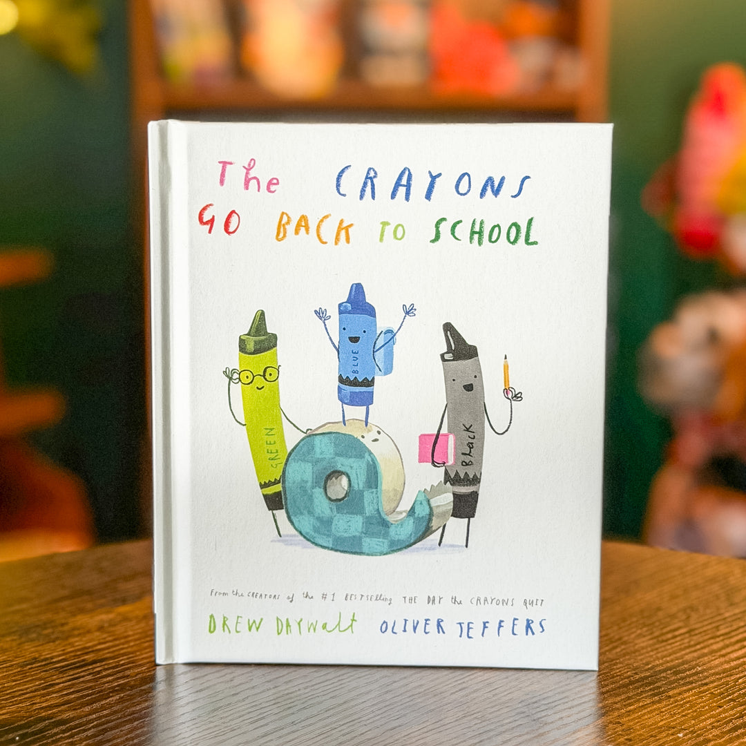 The Crayons Go Back to School book by Drew Daywalt Oliver Teffers, featuring a white cover with colorful hand-drawn crayon characters and a roll of tape, standing on a wooden table with a blurred background.