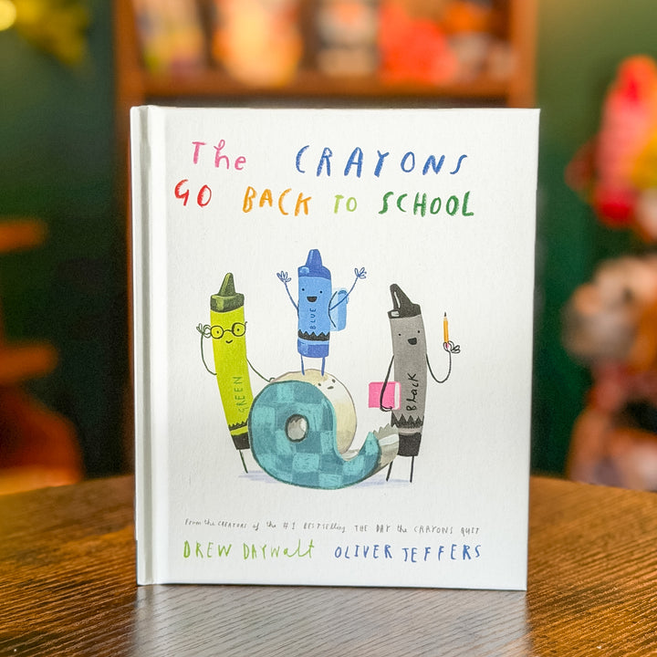 The Crayons Go Back to School book by Drew Daywalt Oliver Teffers, featuring a white cover with colorful hand-drawn crayon characters and a roll of tape, standing on a wooden table with a blurred background.