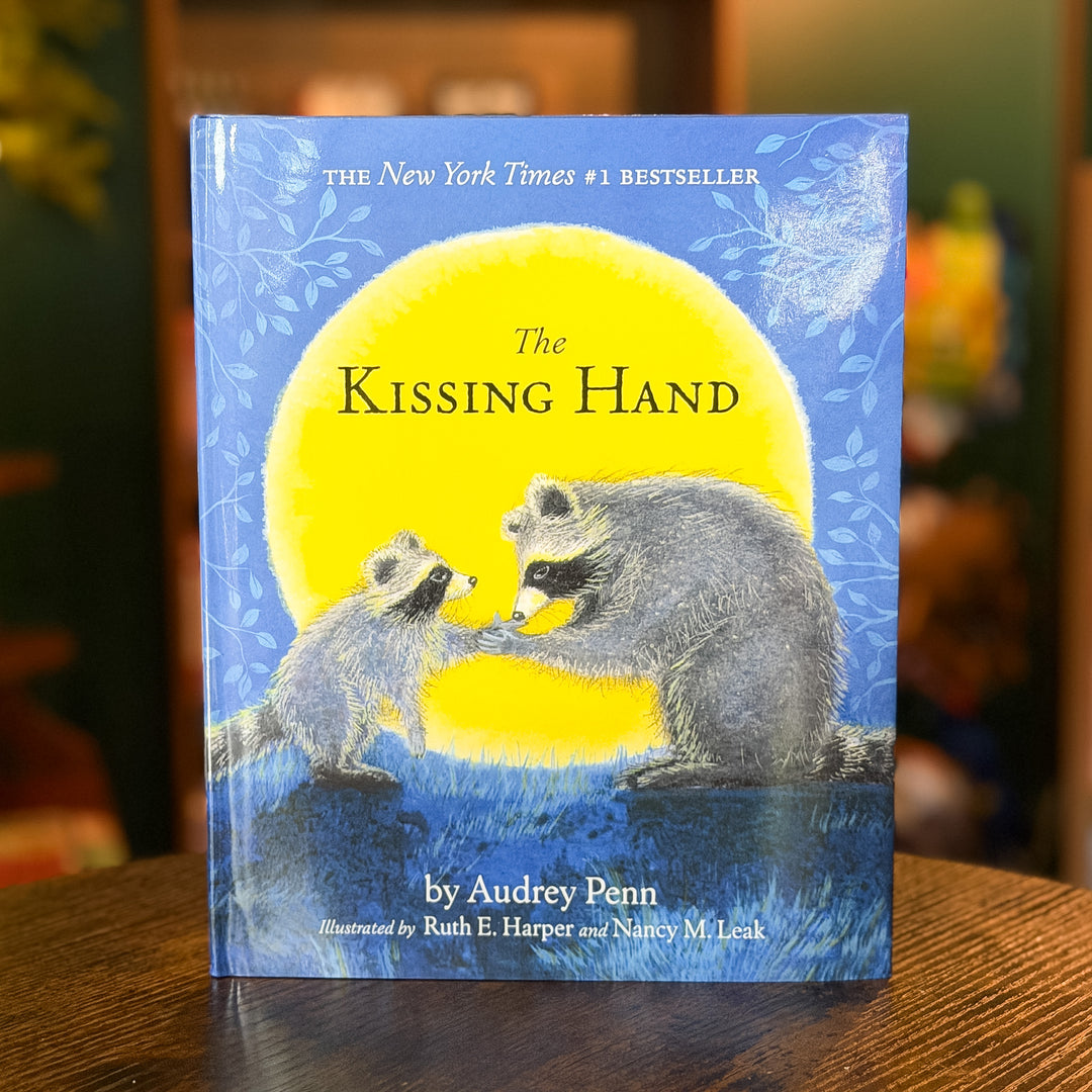 The Kissing Hand book by Audrey Penn, featuring a blue cover with a large yellow moon and an illustration of two raccoons facing each other, displayed on a wooden table with a blurred background.