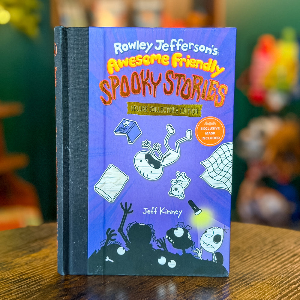 The book 'Rowley Jefferson's Awesome Friendly Spooky Stories' by Jeff Kinney stands upright on a wooden table. The cover is a vibrant purple with various cartoon illustrations of characters and objects, and the title is written in large, playful orange and yellow lettering.