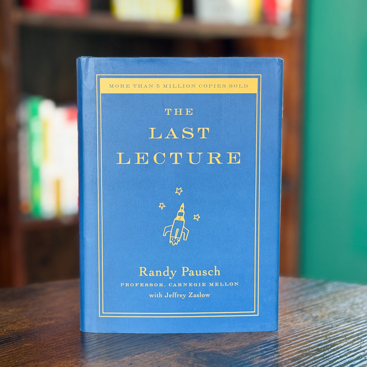 The Last Lecture hardcover book by Randy Pausch, featuring a dusty blue cover with gold lettering and a rocket illustration, standing on a wooden table with a blurred bookshelf in the background.