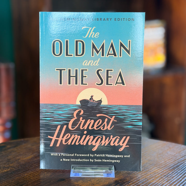 The Old Man and the Sea book cover by Ernest Hemingway, displayed upright on a small clear stand on a wooden table with a blurred indoor background.