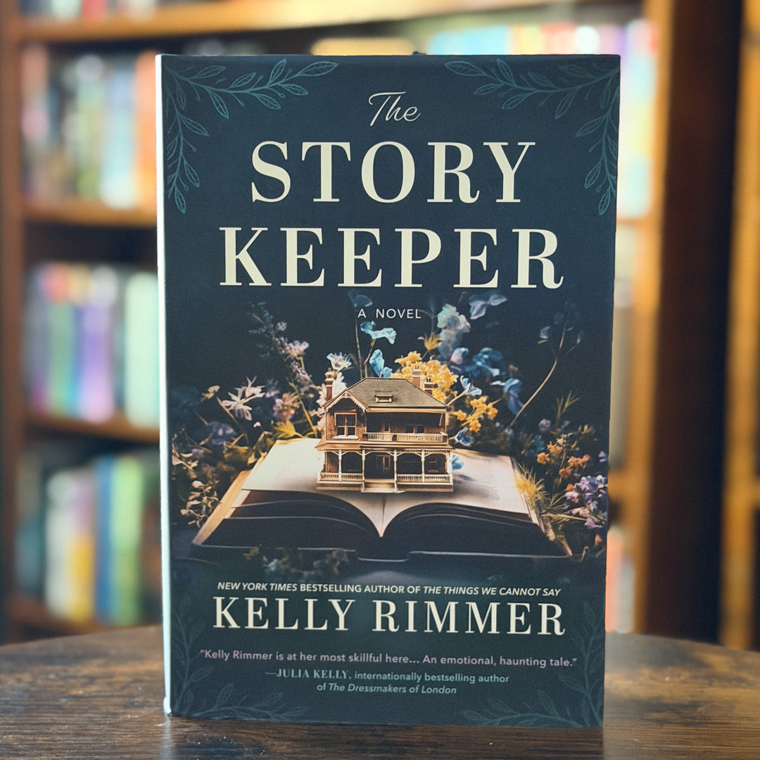 The Story Keeper: A Novel by Kelly Rimmer book cover displayed upright on a wooden table with a blurred library or bookstore background.