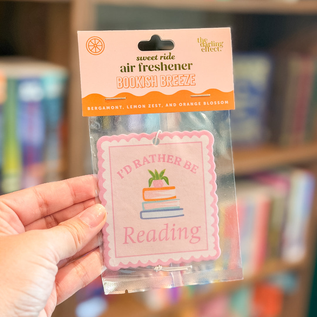 A hand holds a packaged air freshener sticker in a blurred bookstore setting. The product is a square pink sticker with the text 'I'D RATHER BE Reading' and a small illustration of stacked books with a green plant, attached to a peach and orange header card that reads 'sweet ride air freshener BOOKISH BREEZE' and shows the 'The Writing Effect' brand.