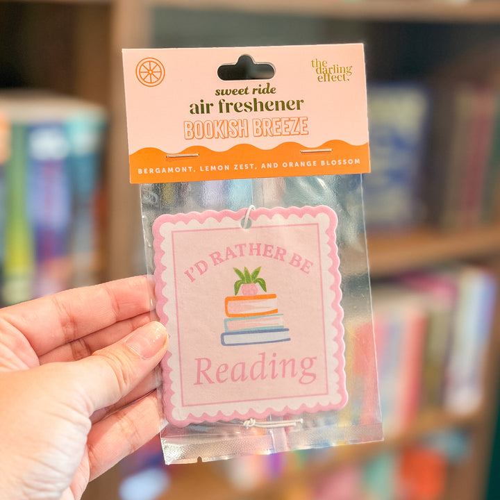 A hand holds a packaged air freshener sticker in a blurred bookstore setting. The product is a square pink sticker with the text &