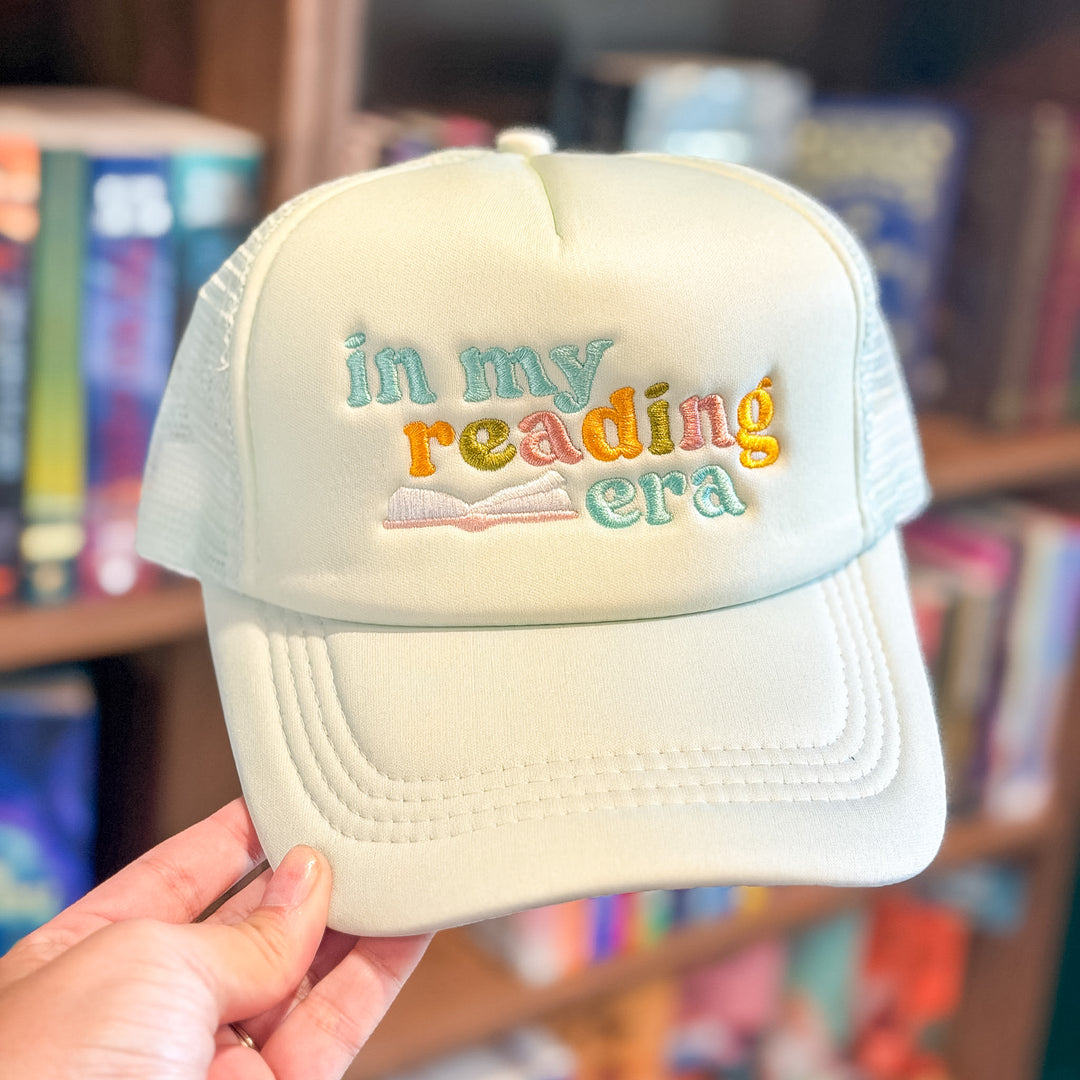 A hand holding a cream-colored trucker hat with colorful embroidered text reading &