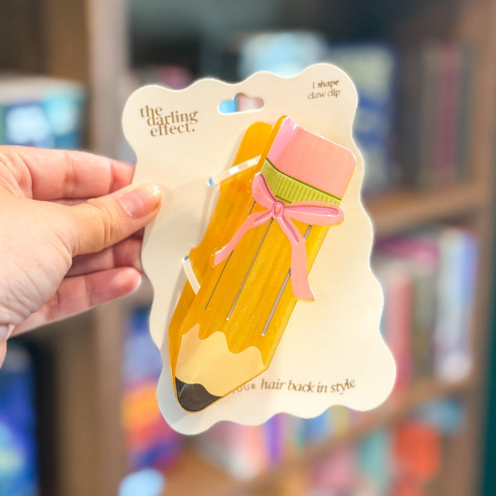 A hand holds a carded hair clip shaped like a yellow pencil with a pink eraser, green center, and pink ribbon bow. The card reads &