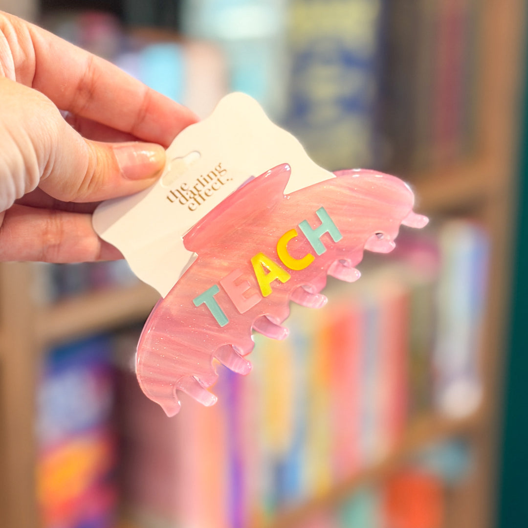 A hand holds a translucent pink hair claw clip with the word 'TEACH' in colorful raised letters on a white backing card. The card shows the brand name 'the fine clothes'.