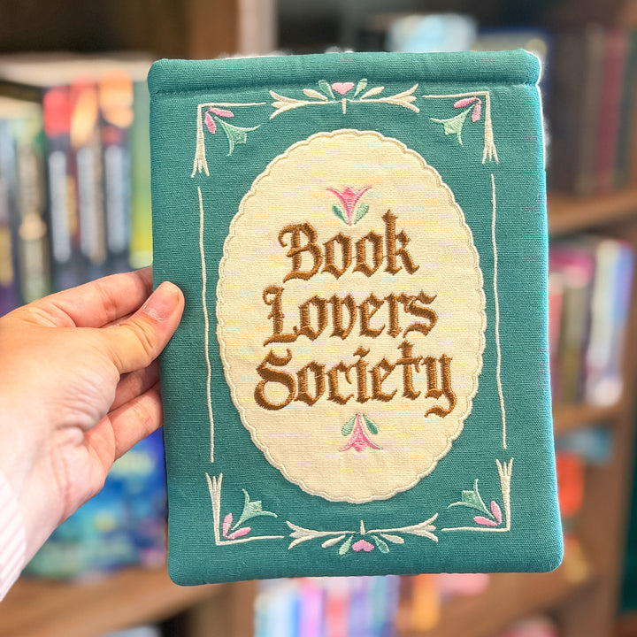 A hand holds a teal embroidered fabric book cover or pouch with a cream oval center reading &