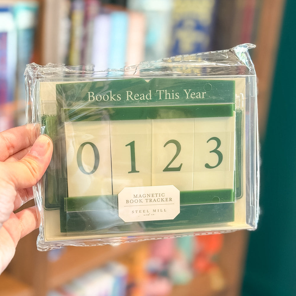 A hand holding a clear plastic-wrapped magnetic book tracker calendar. The tracker shows the numbers 0, 1, 2, 3 in dark green on cream-colored tiles, with a dark green header reading 'Books Read This Year' and a white label reading 'MAGNETIC BOOK TRACKER' and 'STEEL MILL'.