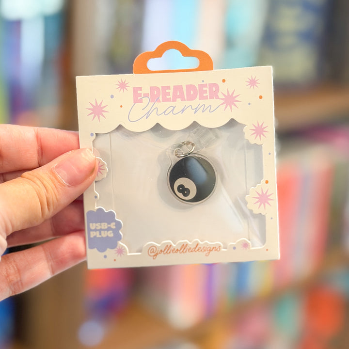 A hand holds a packaged e-reader charm shaped like a black and white 8-ball game piece, mounted on a decorative white card labeled &